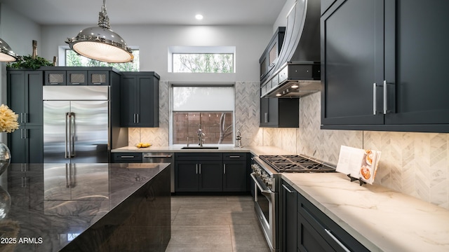 kitchen featuring light stone counters, a sink, high quality appliances, backsplash, and wall chimney exhaust hood