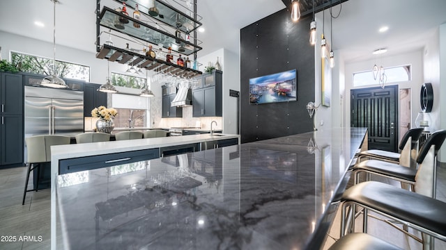 bar with custom range hood, hanging light fixtures, decorative backsplash, a sink, and stainless steel built in refrigerator