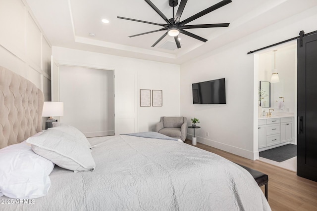 bedroom with ceiling fan, a raised ceiling, a barn door, ensuite bathroom, and light wood-type flooring