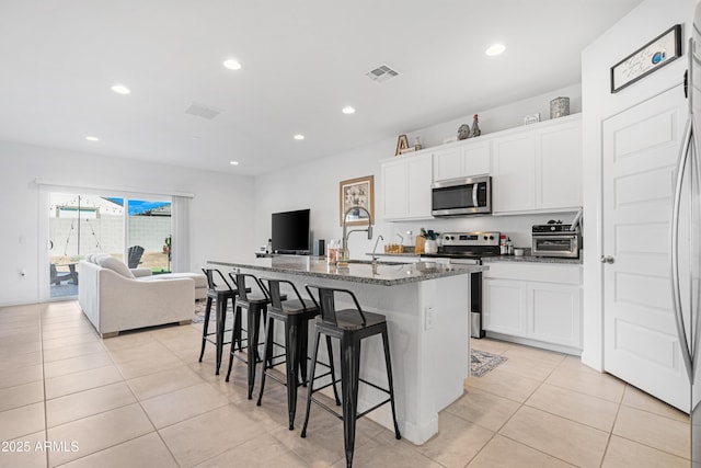 kitchen featuring appliances with stainless steel finishes, dark stone countertops, an island with sink, white cabinets, and light tile patterned flooring