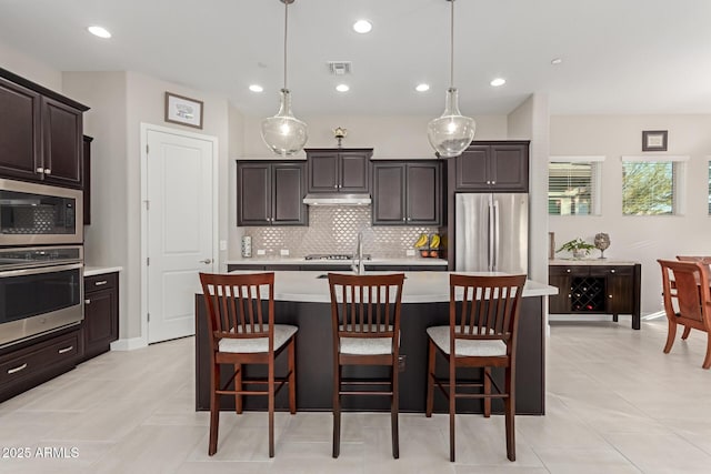 kitchen with decorative light fixtures, dark brown cabinetry, stainless steel appliances, and a kitchen island with sink