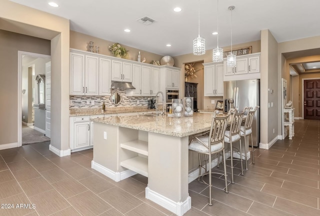 kitchen featuring decorative light fixtures, white cabinetry, an island with sink, light stone countertops, and stainless steel refrigerator with ice dispenser