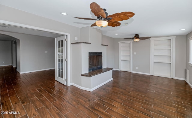 unfurnished living room featuring baseboards, a ceiling fan, a fireplace with raised hearth, arched walkways, and wood finished floors