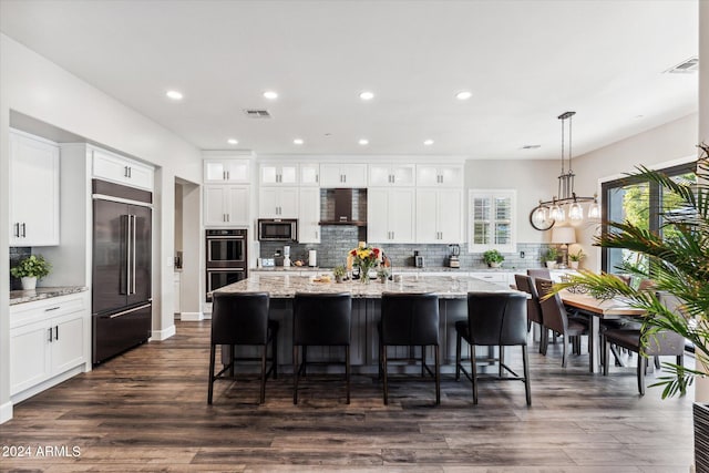 kitchen with wall chimney exhaust hood, dark wood-type flooring, stainless steel appliances, an island with sink, and white cabinets