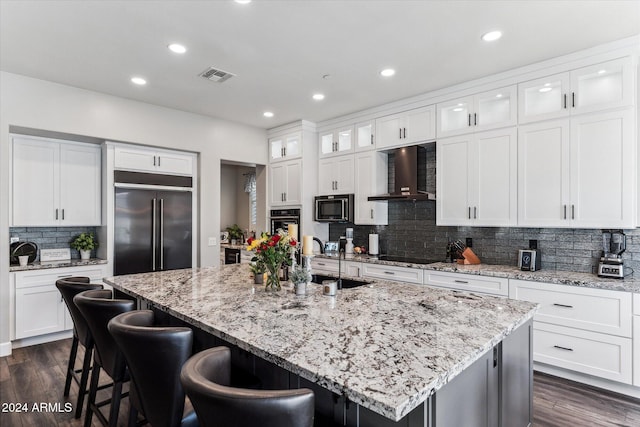 kitchen with wall chimney exhaust hood, built in refrigerator, a center island with sink, and white cabinetry