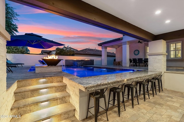 pool at dusk featuring pool water feature, ceiling fan, a patio, and an outdoor bar