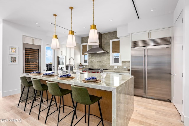 kitchen with light wood-type flooring, tasteful backsplash, an island with sink, stainless steel appliances, and wall chimney exhaust hood