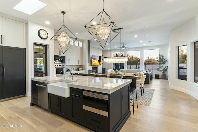 kitchen with an island with sink, ceiling fan, a fireplace, hanging light fixtures, and white cabinets