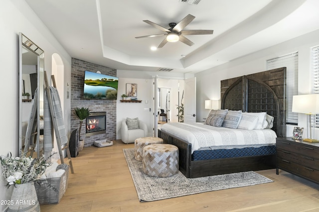 bedroom featuring ceiling fan, a fireplace, a tray ceiling, and light hardwood / wood-style floors