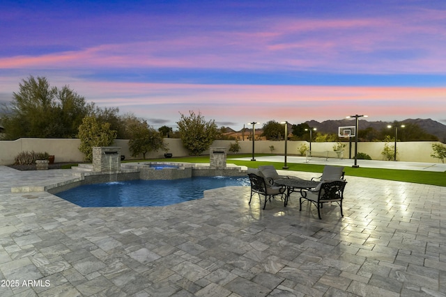 pool at dusk featuring pool water feature, a patio area, and basketball hoop