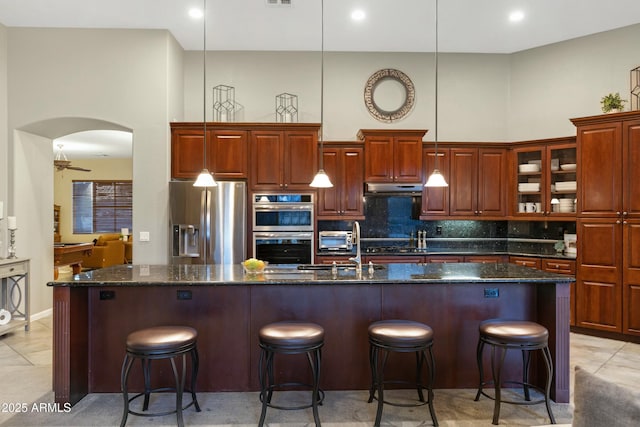 kitchen featuring under cabinet range hood, stainless steel appliances, a towering ceiling, decorative backsplash, and a center island with sink