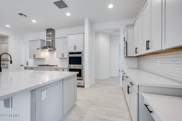 kitchen with light wood-type flooring, tasteful backsplash, light stone counters, wall chimney exhaust hood, and stainless steel appliances