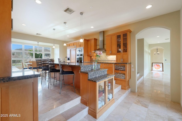 kitchen featuring built in appliances, wall chimney exhaust hood, dark stone countertops, an island with sink, and decorative light fixtures