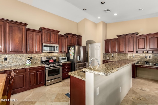 kitchen with visible vents, a towering ceiling, light stone counters, a kitchen island with sink, and stainless steel appliances