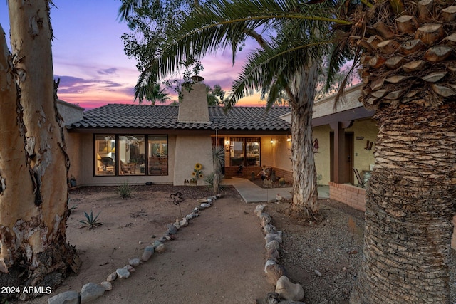 back of house at dusk with a tiled roof, a chimney, and stucco siding