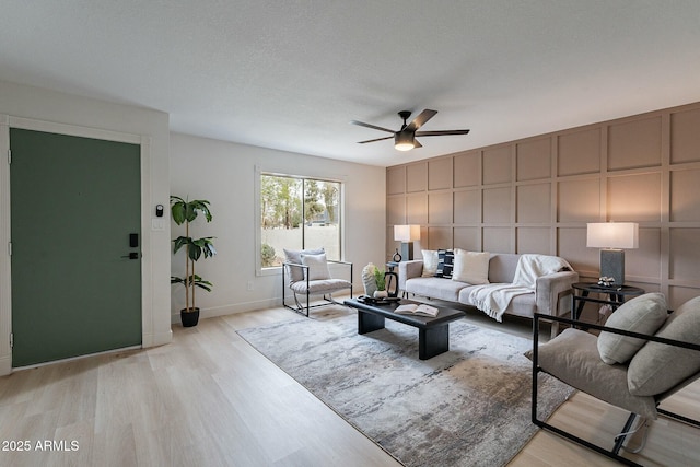 living area with light wood-type flooring, a ceiling fan, a textured ceiling, a decorative wall, and baseboards