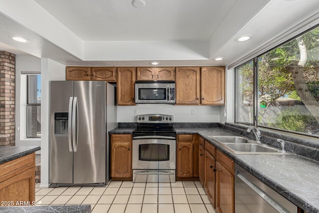 kitchen with dark countertops, appliances with stainless steel finishes, a sink, brown cabinetry, and light tile patterned floors