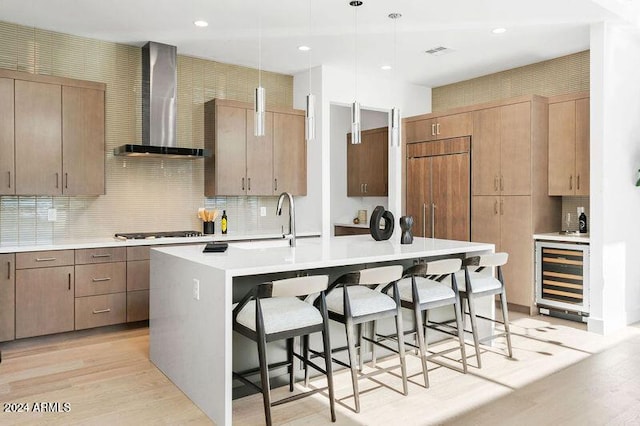 kitchen with wine cooler, decorative light fixtures, a kitchen island with sink, wall chimney range hood, and light wood-type flooring