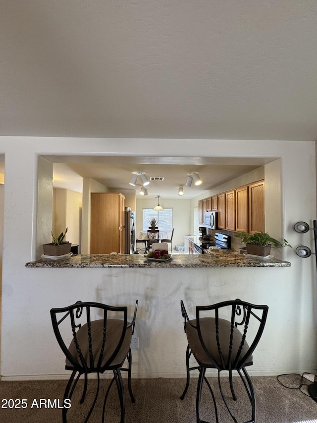 kitchen with a breakfast bar, brown cabinets, stainless steel appliances, dark stone counters, and a peninsula