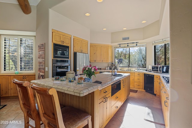 kitchen with beam ceiling, tile countertops, a kitchen island with sink, light tile patterned floors, and black appliances