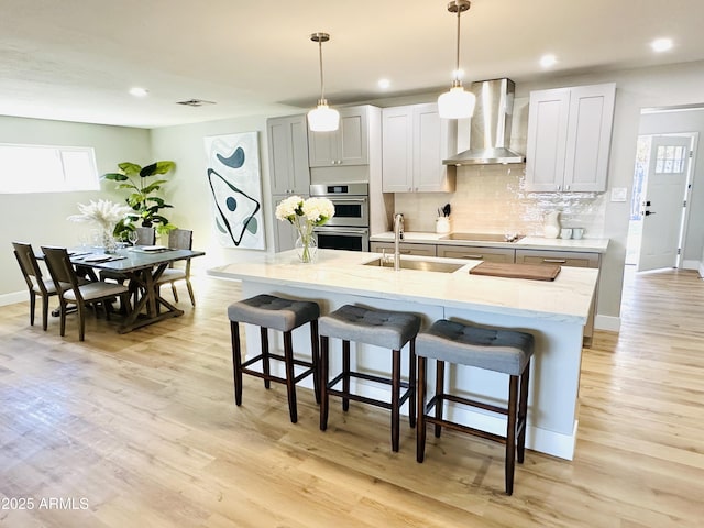 kitchen with a breakfast bar area, a sink, decorative backsplash, double oven, and wall chimney exhaust hood