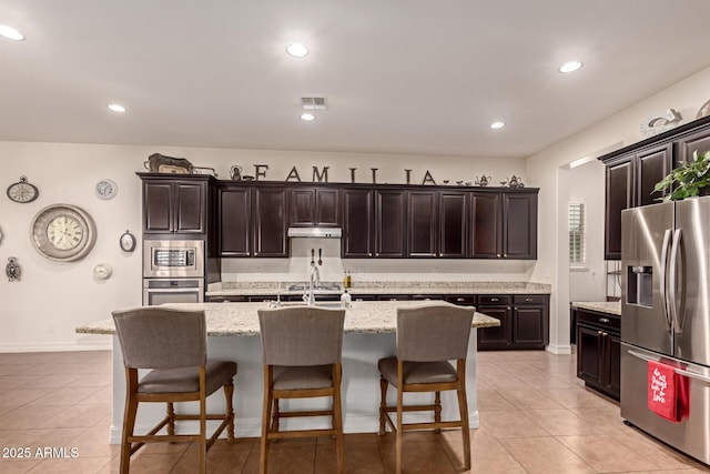 kitchen featuring light tile patterned floors, appliances with stainless steel finishes, an island with sink, and dark brown cabinetry