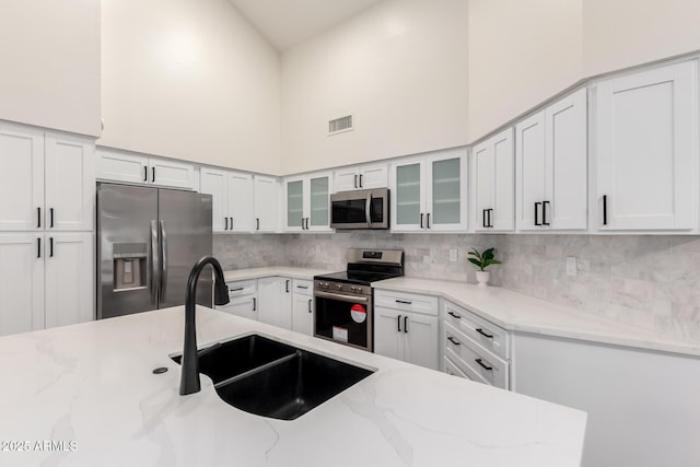 kitchen with appliances with stainless steel finishes, a sink, a towering ceiling, tasteful backsplash, and white cabinetry