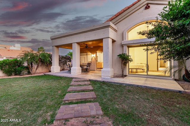 rear view of house with ceiling fan, a lawn, a patio, and stucco siding