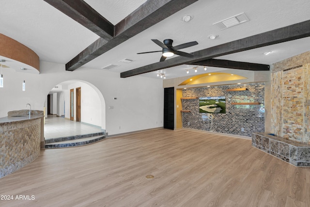 unfurnished living room featuring a textured ceiling, wood-type flooring, ceiling fan, brick wall, and beam ceiling