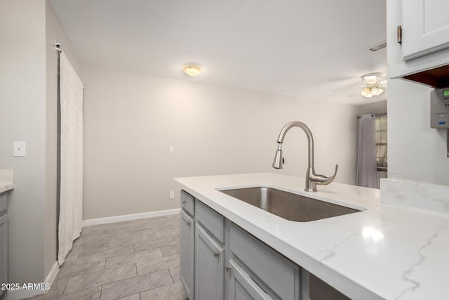 kitchen featuring a ceiling fan, gray cabinets, a sink, baseboards, and light stone countertops