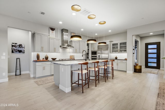 kitchen with a kitchen island with sink, wall chimney exhaust hood, pendant lighting, light hardwood / wood-style flooring, and built in appliances