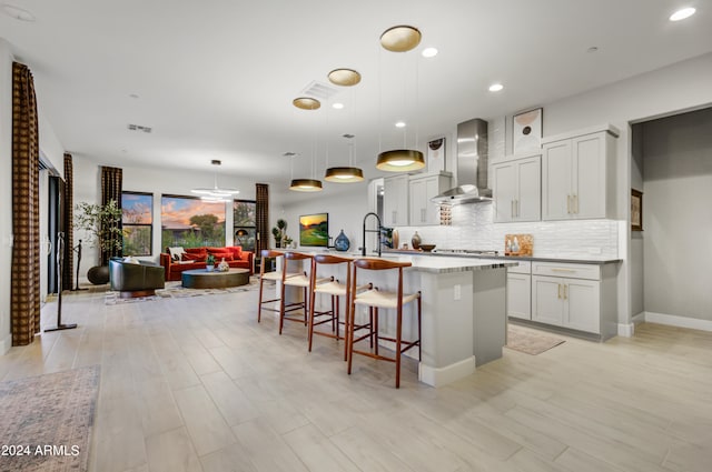 kitchen with a kitchen island with sink, white cabinets, wall chimney exhaust hood, hanging light fixtures, and a breakfast bar area