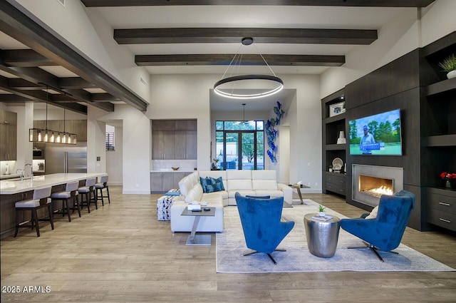living room featuring a notable chandelier, beam ceiling, light wood finished floors, baseboards, and a lit fireplace