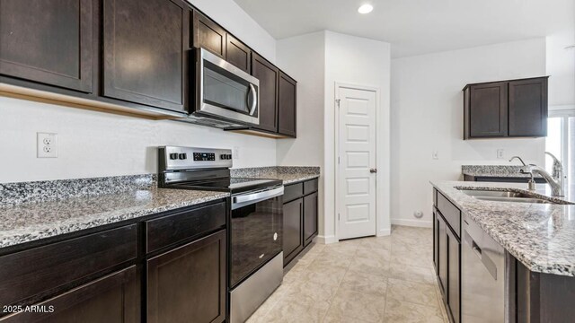 kitchen with light stone counters, stainless steel appliances, a sink, dark brown cabinets, and baseboards