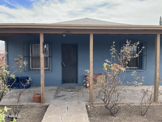 entrance to property featuring a shingled roof, covered porch, and stucco siding