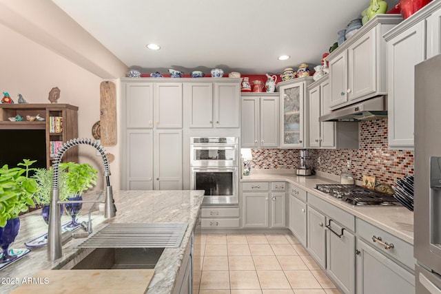 kitchen with under cabinet range hood, stainless steel appliances, a sink, glass insert cabinets, and tasteful backsplash