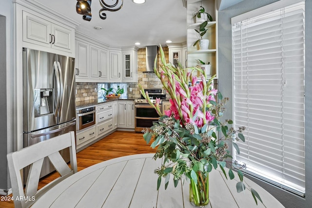 kitchen featuring appliances with stainless steel finishes, wall chimney exhaust hood, white cabinets, light wood-type flooring, and decorative backsplash