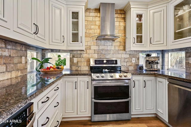 kitchen featuring stainless steel appliances, wall chimney range hood, backsplash, light wood-type flooring, and dark stone countertops