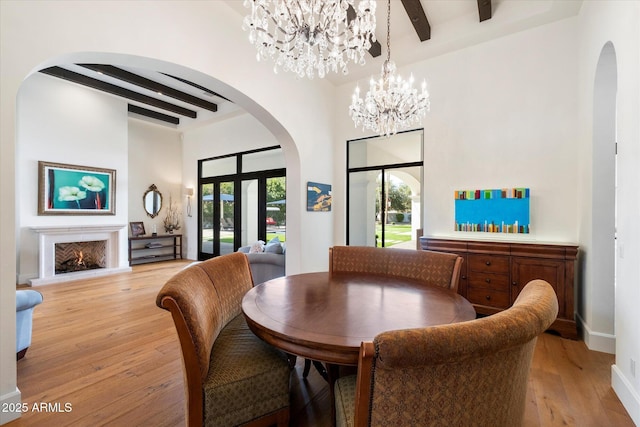 dining room featuring arched walkways, beamed ceiling, a lit fireplace, light wood-type flooring, and a notable chandelier