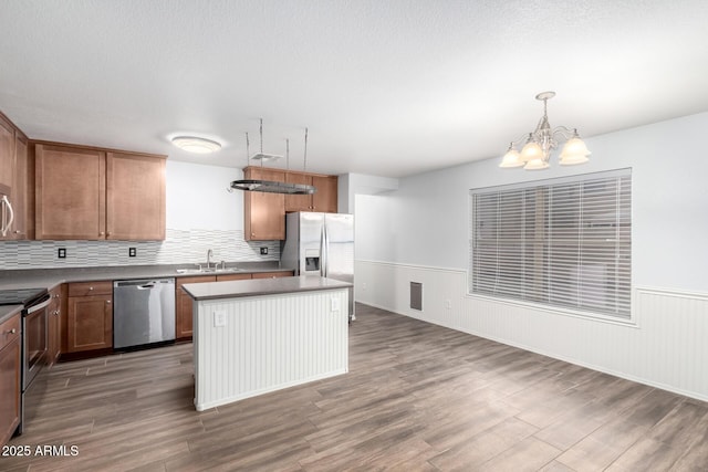 kitchen featuring a wainscoted wall, a kitchen island, wood finished floors, appliances with stainless steel finishes, and decorative light fixtures