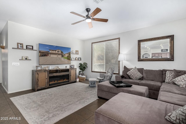 living area featuring a ceiling fan, a glass covered fireplace, dark tile patterned floors, and baseboards