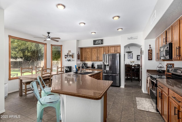 kitchen featuring arched walkways, a breakfast bar area, appliances with stainless steel finishes, brown cabinets, and a center island with sink