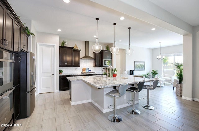 kitchen with wall chimney exhaust hood, light stone counters, hanging light fixtures, a kitchen breakfast bar, and a kitchen island with sink