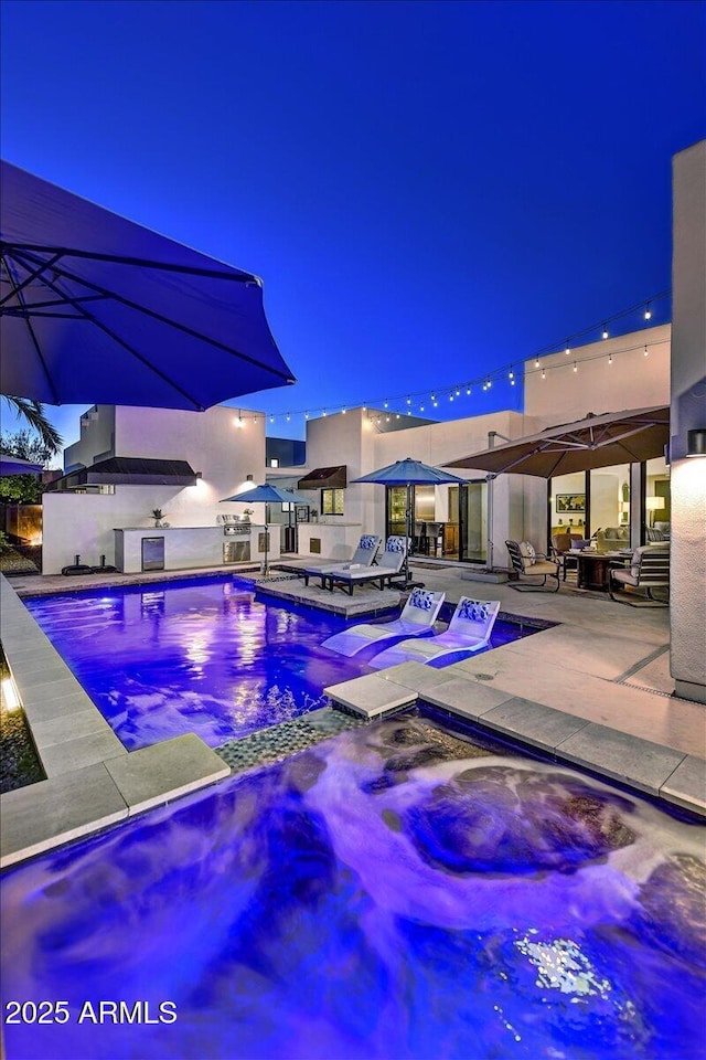 pool at night with exterior kitchen, a jacuzzi, a patio area, and an outdoor pool