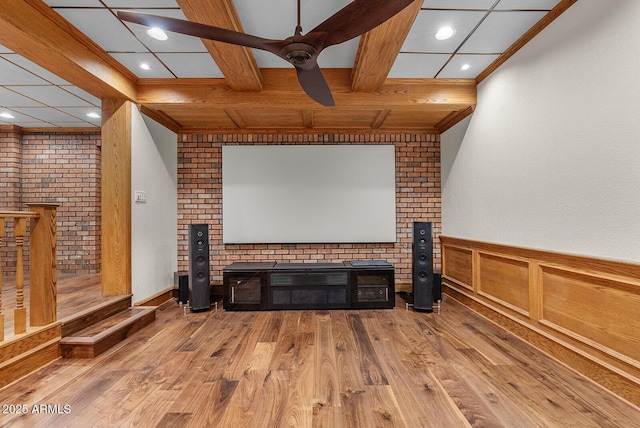 home theater room featuring a ceiling fan, wainscoting, wood finished floors, crown molding, and beam ceiling