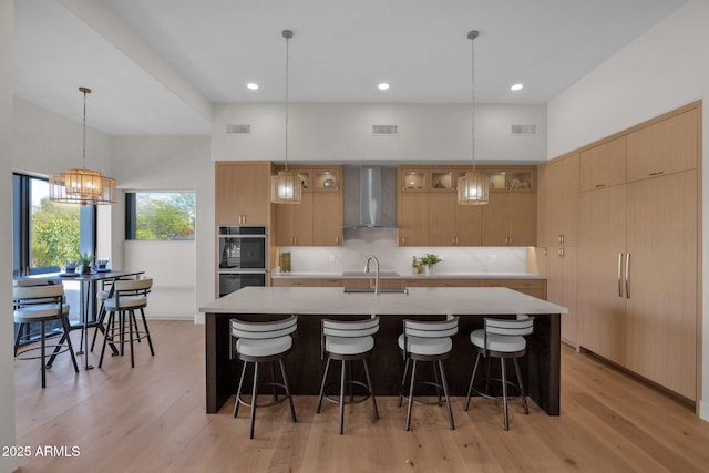 kitchen featuring hanging light fixtures, an island with sink, black double oven, light hardwood / wood-style floors, and wall chimney range hood