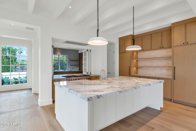 kitchen with beam ceiling, light stone counters, light hardwood / wood-style floors, decorative light fixtures, and a kitchen island with sink