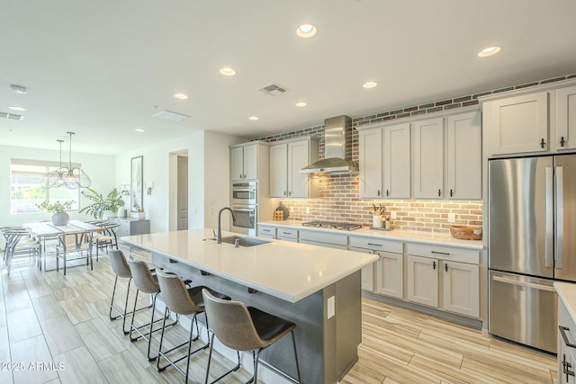 kitchen with pendant lighting, a kitchen island with sink, wall chimney range hood, sink, and stainless steel appliances