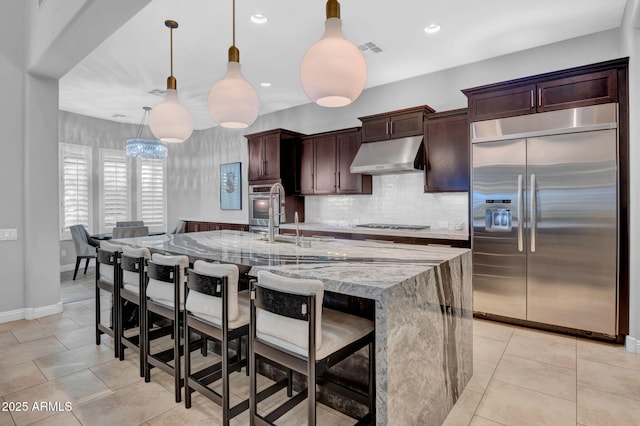 kitchen featuring built in fridge, hanging light fixtures, a kitchen island with sink, and light stone countertops