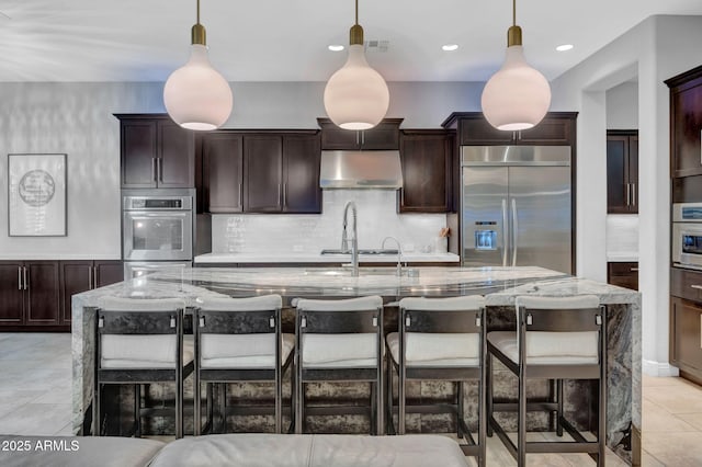 kitchen featuring light stone counters, hanging light fixtures, stainless steel built in fridge, and a large island with sink